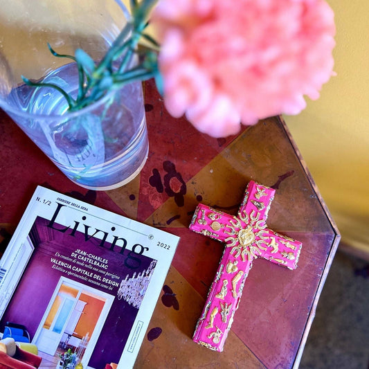 Croix mexicaine rose faite main avec porte-bonheur dorés appelés Milagros dans un décor ethnique et bohème.